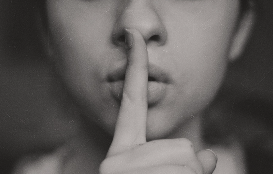Black-and-white close-up of a person holding a finger to their lips in a ‘shh’ gesture.
